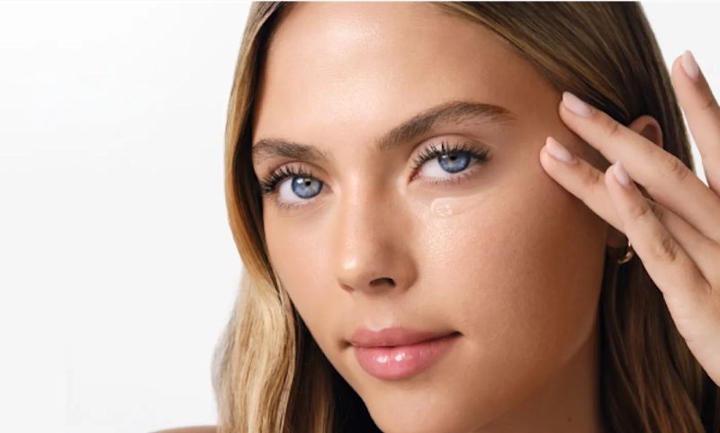 A young woman is applying a clear gel or cream to the skin under her right eye with her fingers.