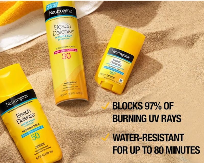  A flat lay shot of three Neutrogena Beach Defense products—a sunscreen lotion, a sunscreen spray, and a sunblock stick—arranged on a towel over sand.