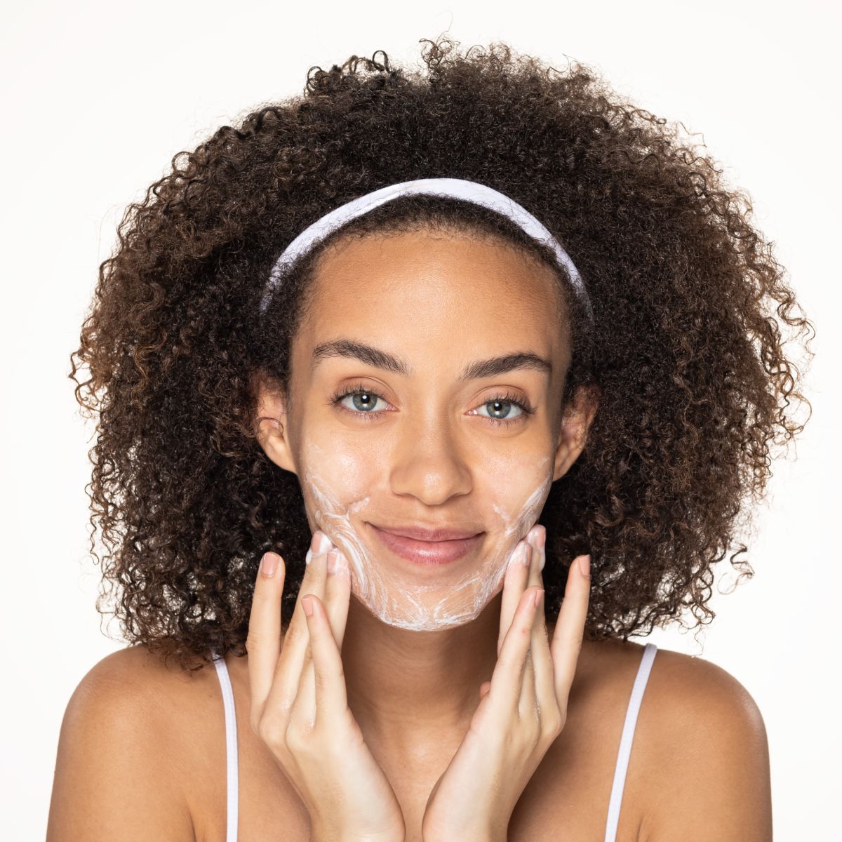 A smiling person with curly hair and a white headband massaging a white creamy cleanser into their face.
