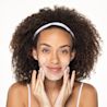 A smiling person with curly hair and a white headband massaging a white creamy cleanser into their face.