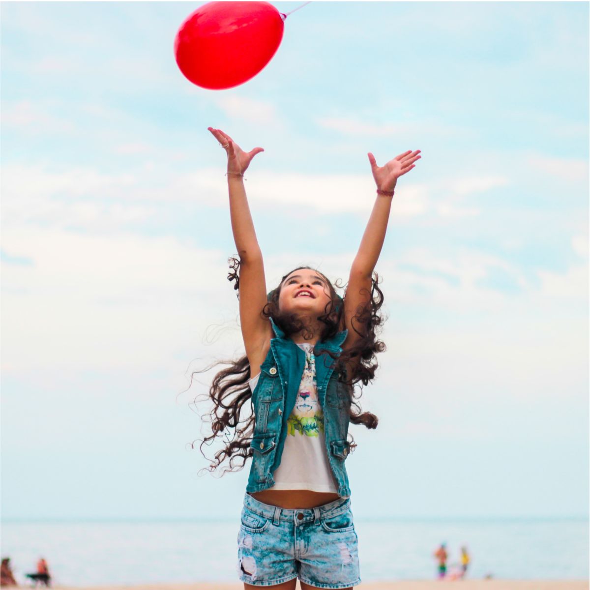 A young girl on a beach reaches up toward a red balloon floating in the sky.