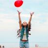 A young girl on a beach reaches up toward a red balloon floating in the sky.