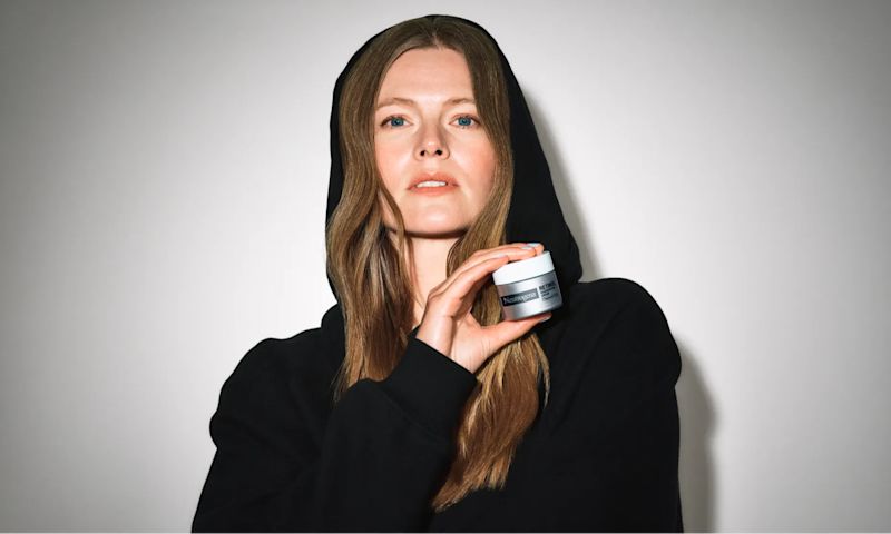 Woman with clear skin wearing a hoodie and holding a jar of Neutrogena® Rapid Wrinkle Repair® Retinol Regenerating Cream Fragrance Free