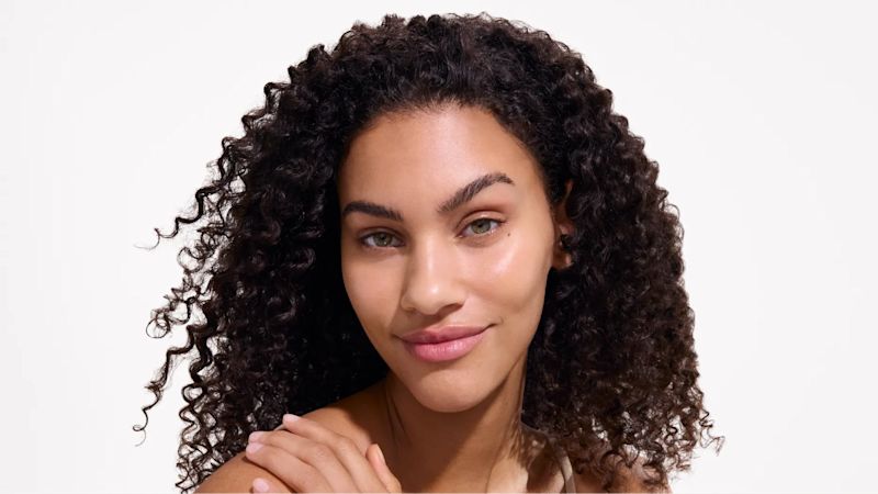 Close-up of a woman with curly hair and natural makeup, smiling softly with glowing skin after applying Neutrogena® Ultra Sheer® Sunscreen