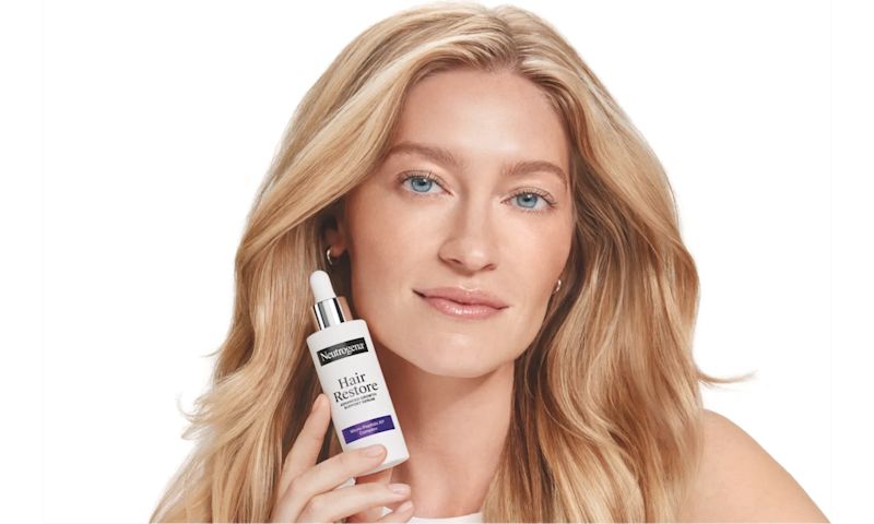 A blonde woman with long, wavy hair holding a Neutrogena Hair Restore serum bottle.