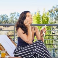 Woman sitting outdoors on a lounge chair, applying skincare product to her face.