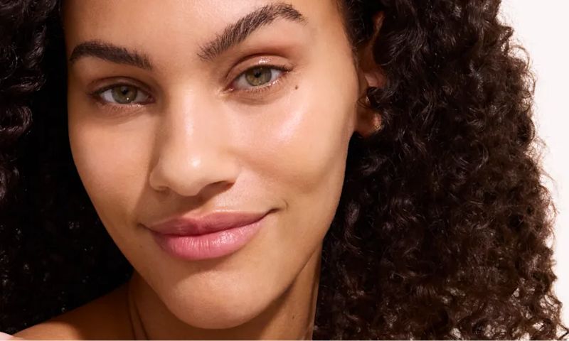 Close-up of a woman with curly hair and natural makeup, smiling softly with glowing skin against a neutral background.