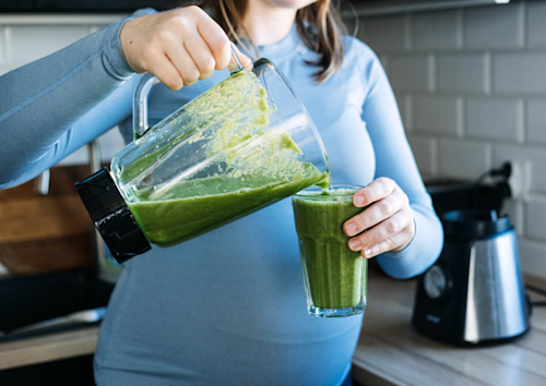 Una mujer está sirviendo en un vaso una bebida saludable de color verde que viene de la batidora.