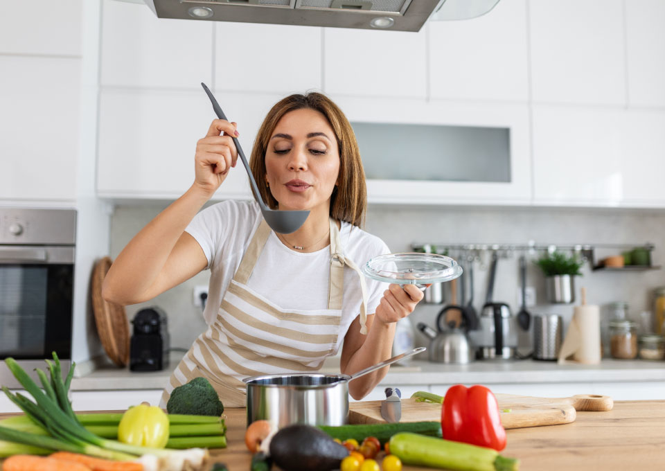 Una mujer está en la cocina soplando sobre la comida caliente que tiene en una cuchara. 