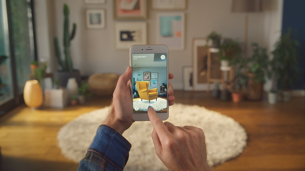 Hands hold up a cell phone to face a living room as they use the Ikea app’s real-time augmented reality feature.