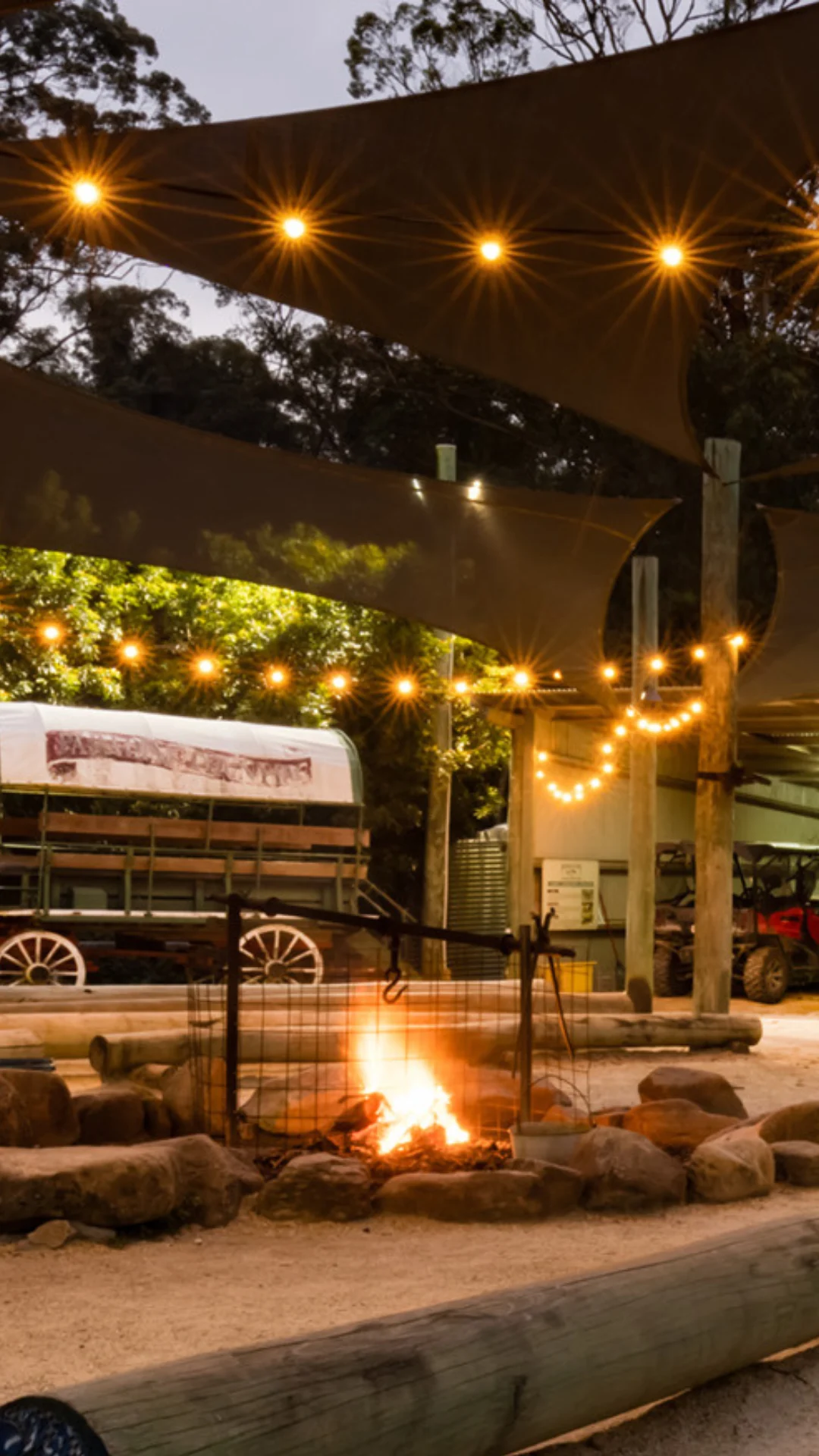 A campfire burns within a stone ring, surrounded by string lights and a covered seating area; a covered wagon is visible in the background.