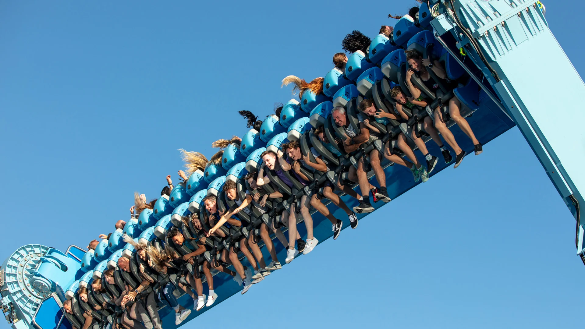 People ride a blue amusement park thrill ride, suspended upside down against a clear blue sky, with hair and arms flung by the motion.