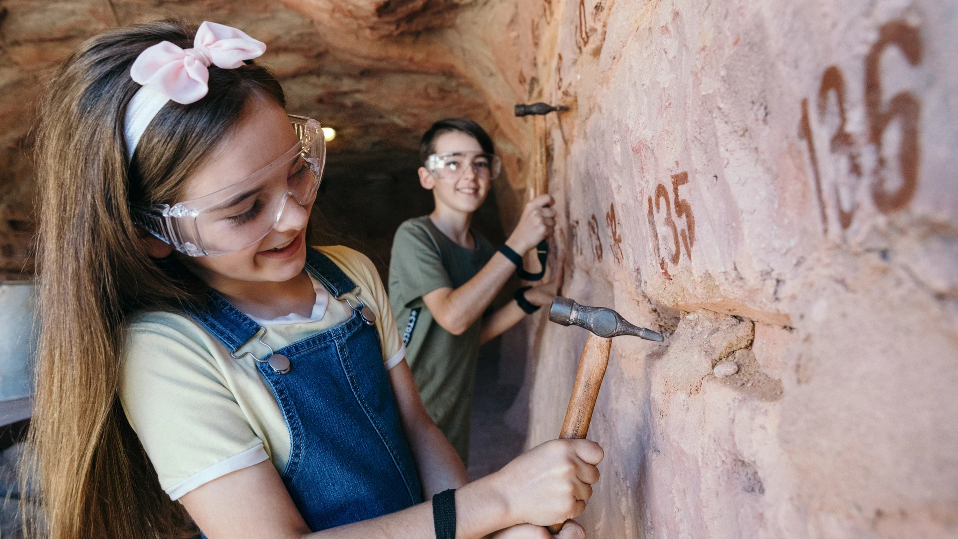 Two children wearing safety goggles use chisels and hammers to work on a numbered rock wall inside a cave-like setting.