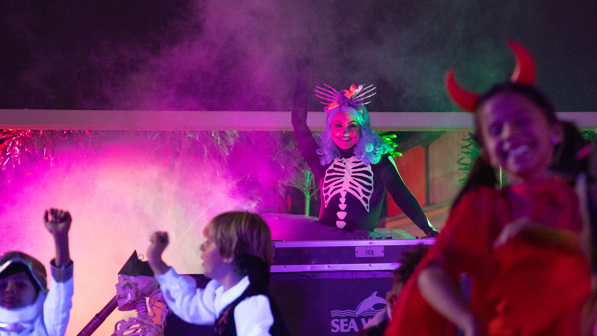 Children in Halloween costumes dance energetically. In the background, a person dressed in a skeleton costume with silver hair and a dramatic headdress stands with one arm raised, surrounded by colorful fog and lights.