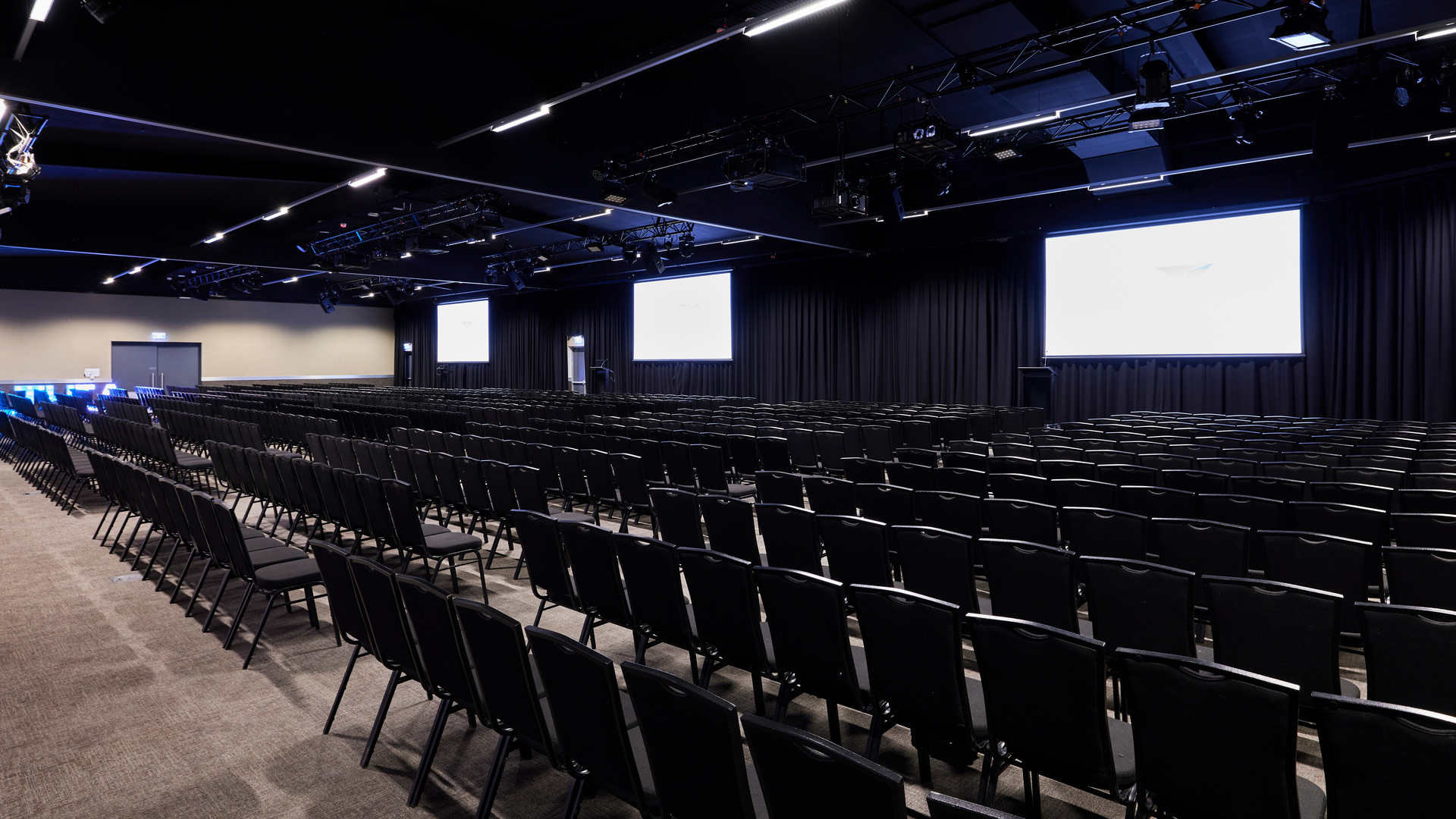 A large, empty conference room filled with rows of black chairs facing several projection screens and a stage, with dim lighting and dark curtains lining the walls.