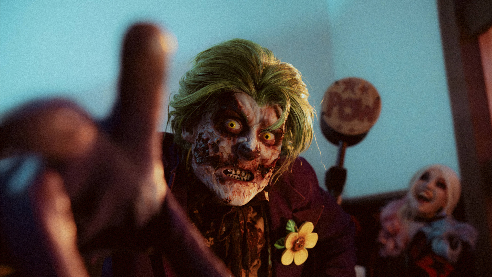 A person in a creepy zombie clown costume with green hair and glowing eyes reaches toward the camera. Another person in clown makeup and costume smiles in the background. The scene is dimly lit, creating a spooky atmosphere.