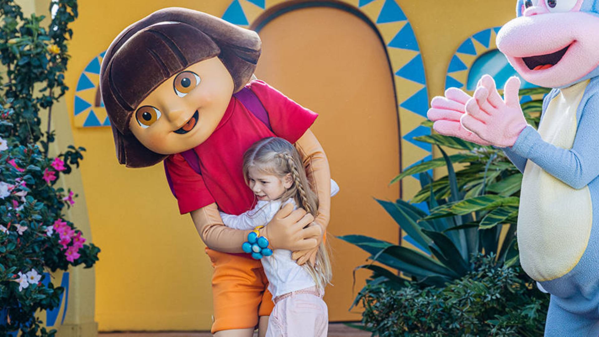 A child smiles while hugging a person in a Dora the Explorer costume outdoors, with colorful plants and another character in the background.