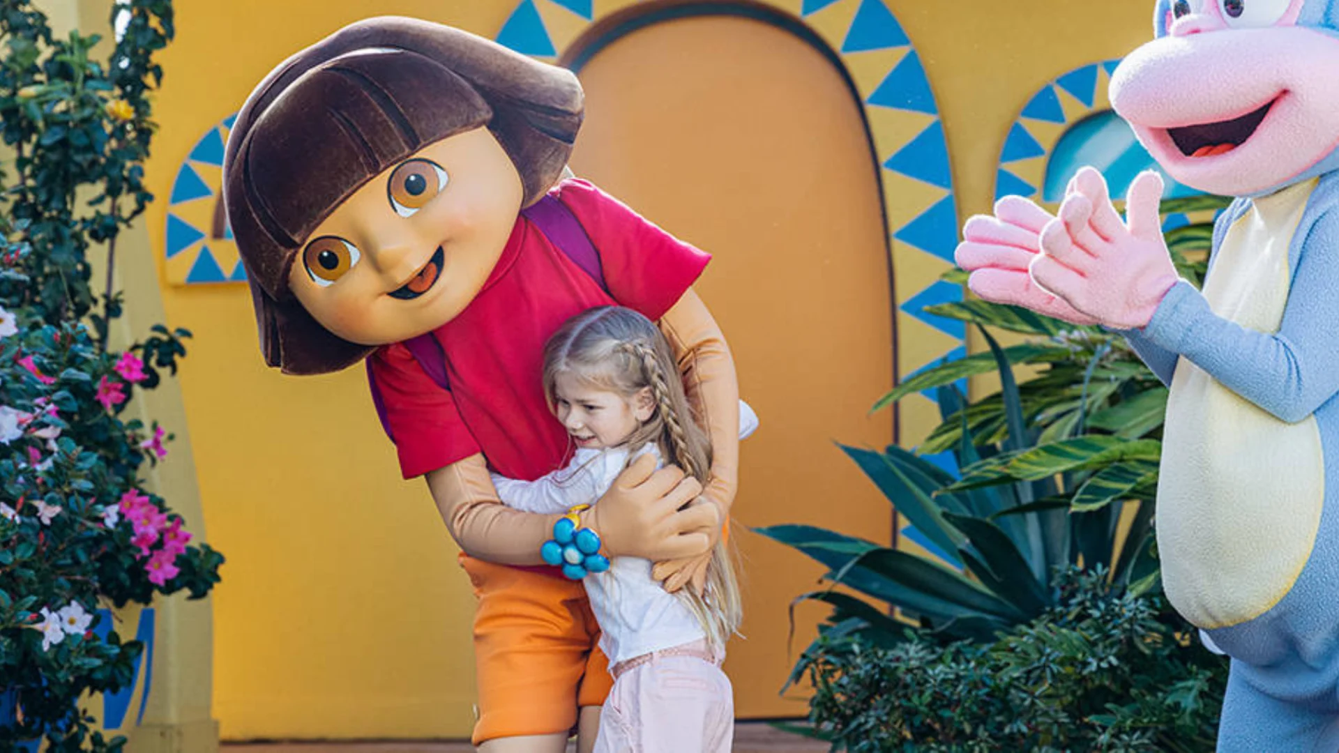 A child smiles while hugging a person in a Dora the Explorer costume outdoors, with colorful plants and another character in the background.