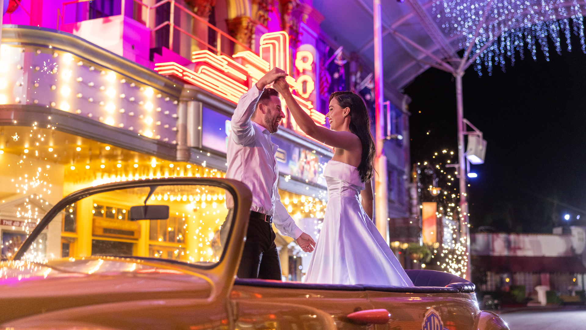 A couple dressed in formal attire dances in the back of a classic convertible car at night, surrounded by colorful neon lights and festive decorations outside a lively, retro-style building.