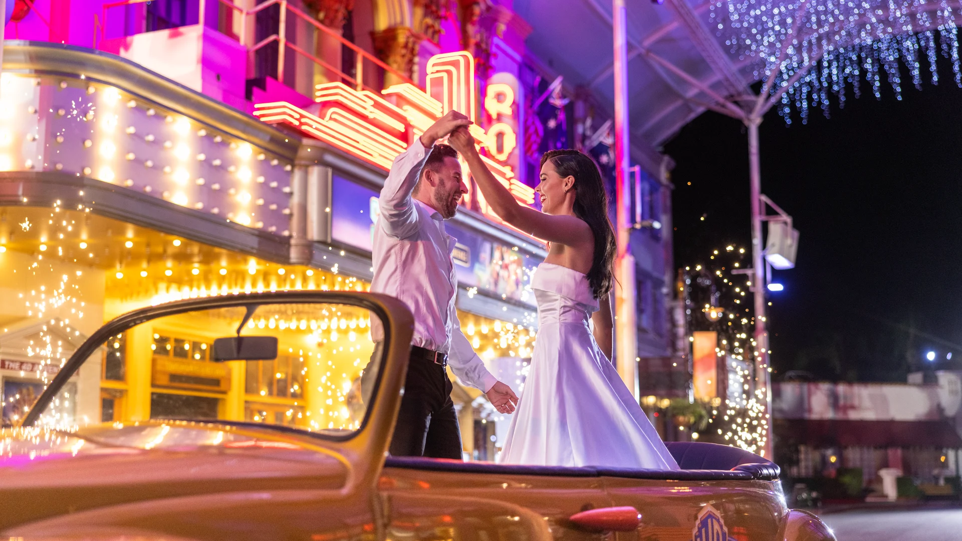 A couple dressed in formal attire dances in the back of a classic convertible car at night, surrounded by colorful neon lights and festive decorations outside a lively, retro-style building.
