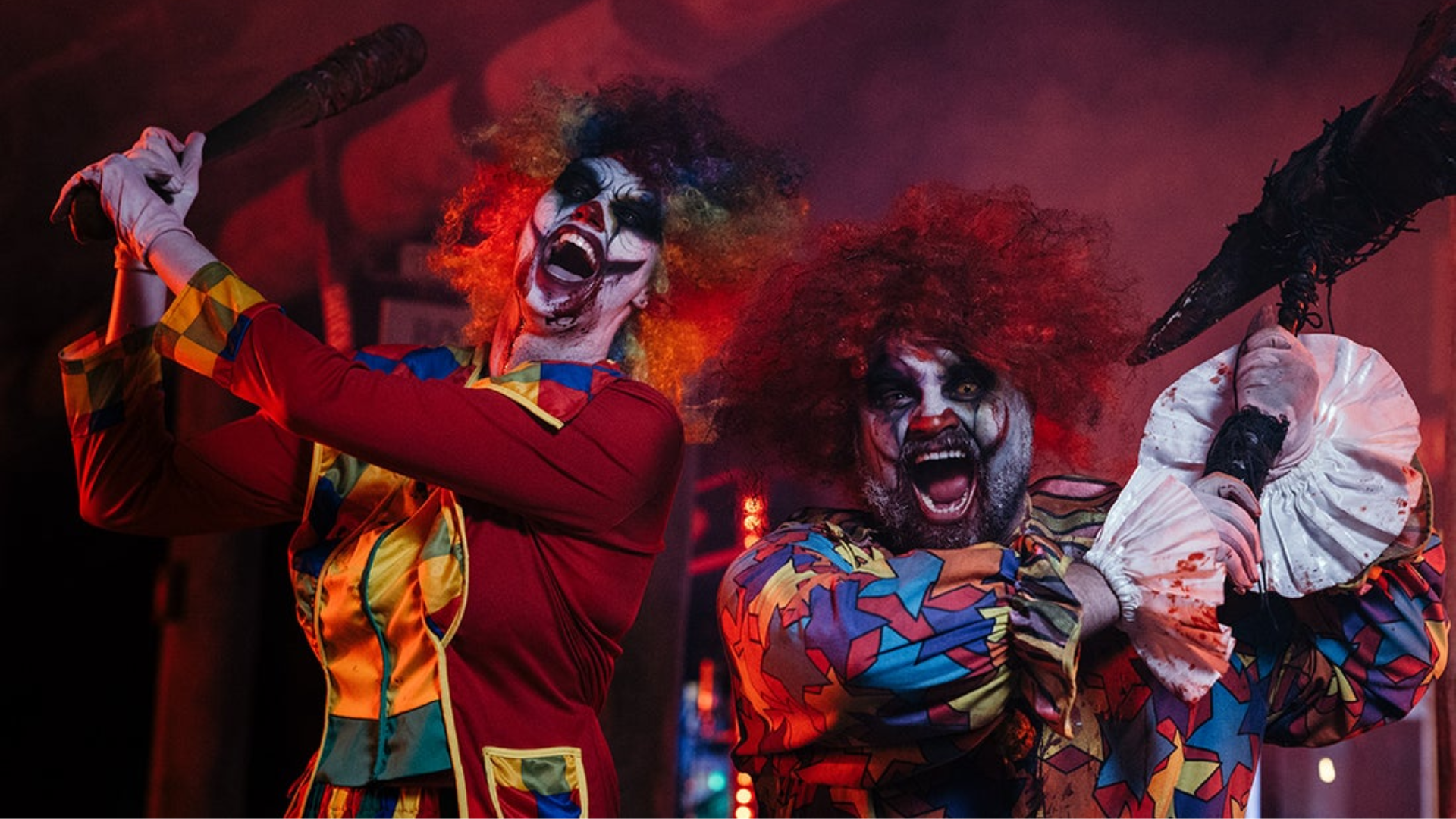 Two people dressed as scary clowns with colorful wigs and face paint, holding large spiked clubs, pose menacingly in dramatic, dark lighting with smoke in the background.