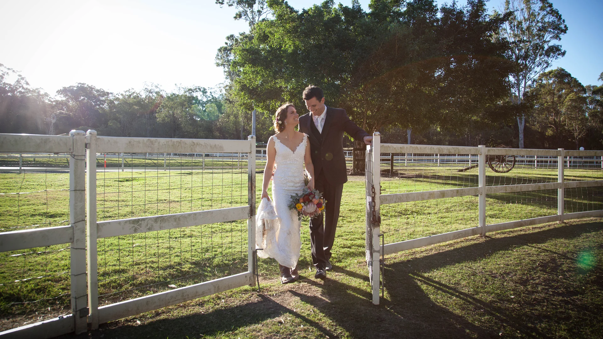 A bride in a white dress and a groom in a dark suit walk together by a white fence on a sunny day, smiling and holding hands, with green trees and grass in the background.