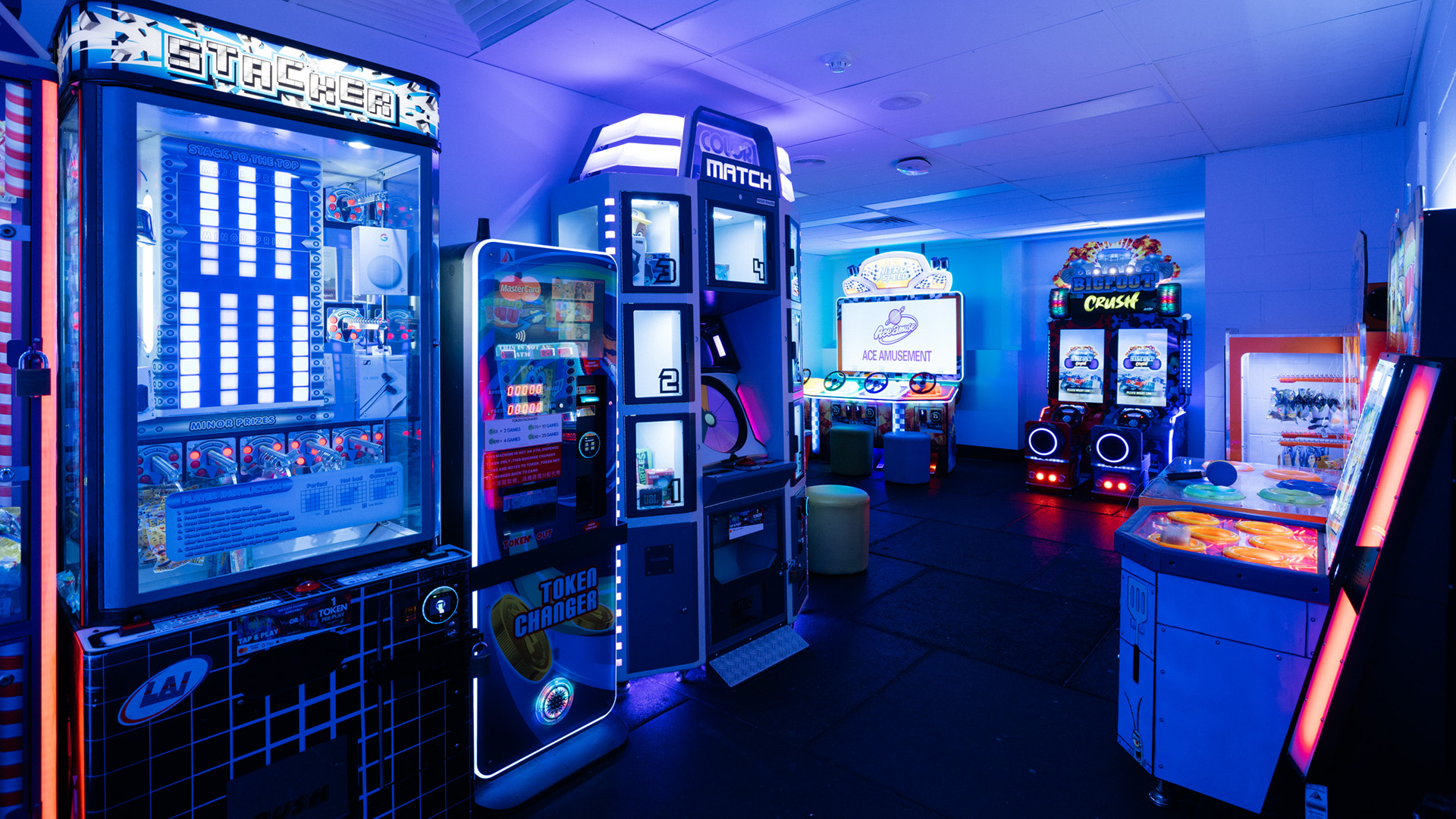 A neon-lit arcade with various game machines, including stackers, a dance game, and a driving game setup, set in a dimly lit room.