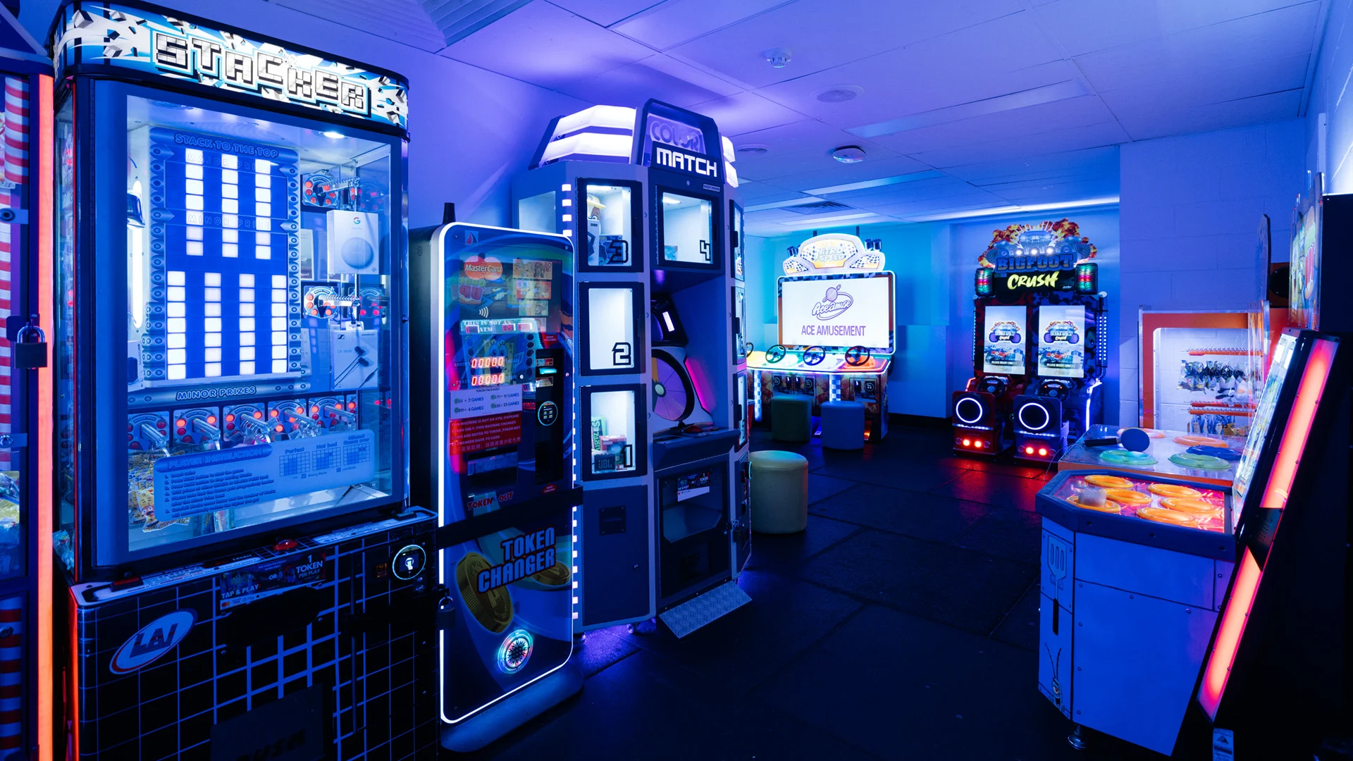 A neon-lit arcade with various game machines, including stackers, a dance game, and a driving game setup, set in a dimly lit room.
