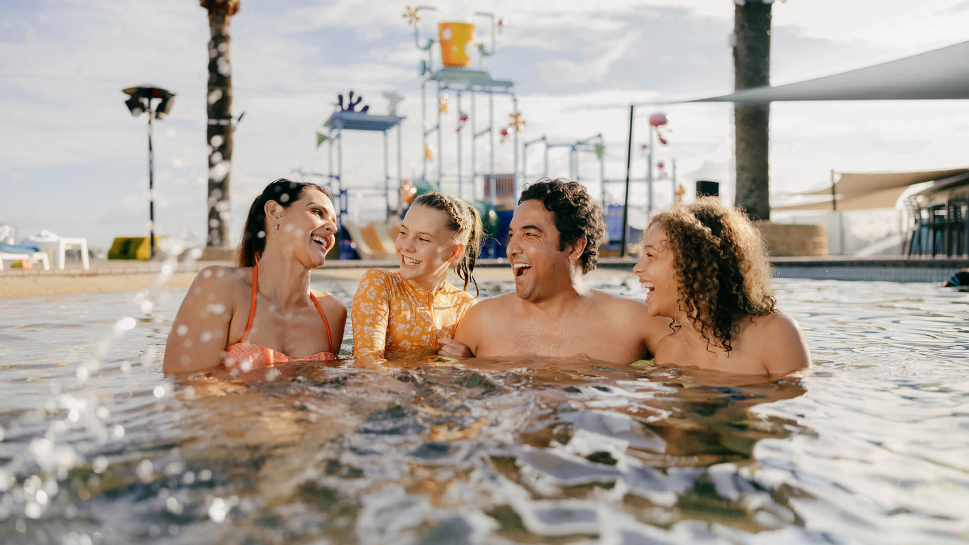 Four people smile and chat while standing in a pool, with a water park structure visible in the background.