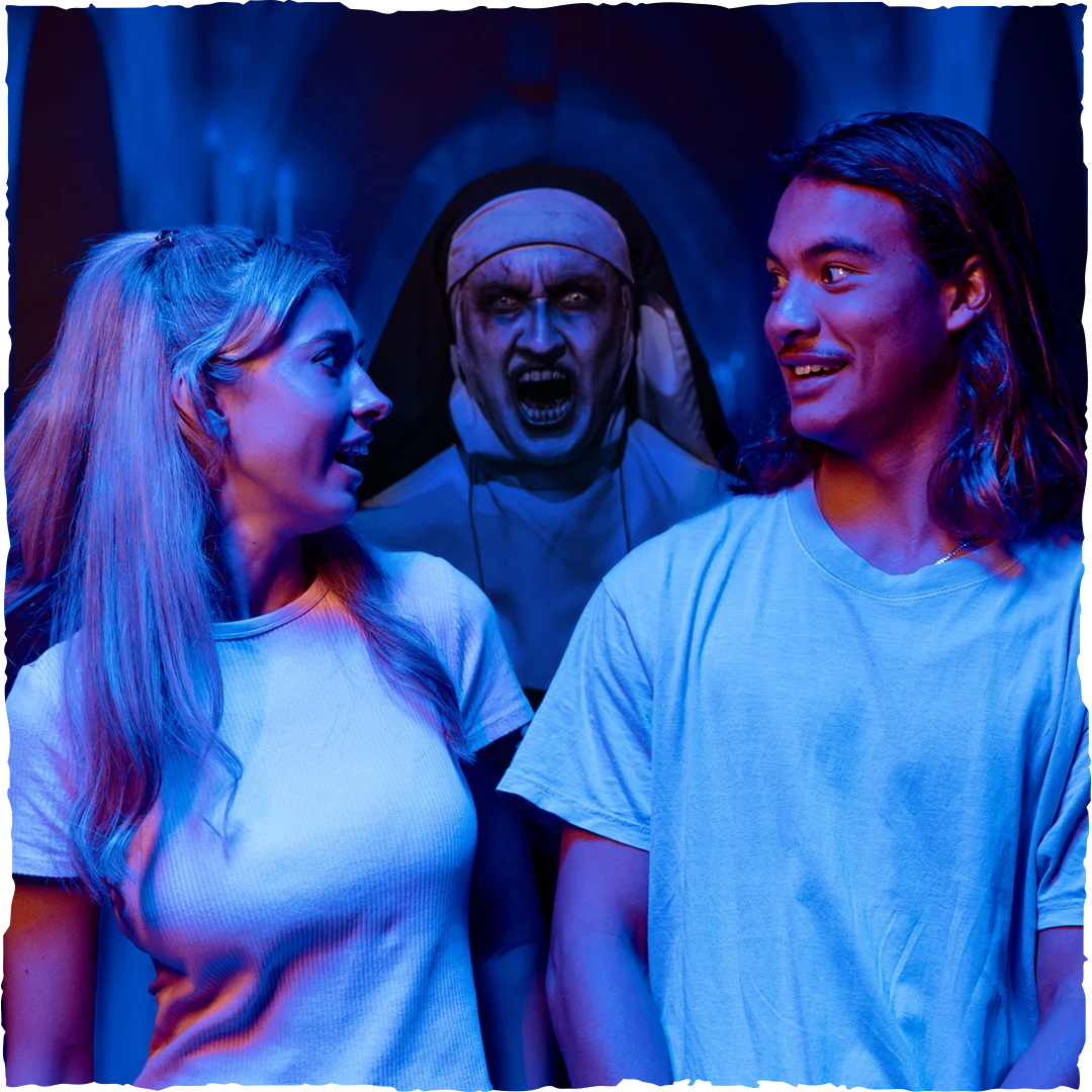A young woman and man, both smiling and surprised, are illuminated by blue and pink lights. Behind them, a person dressed as a scary nun with a menacing expression lurks in the shadows.