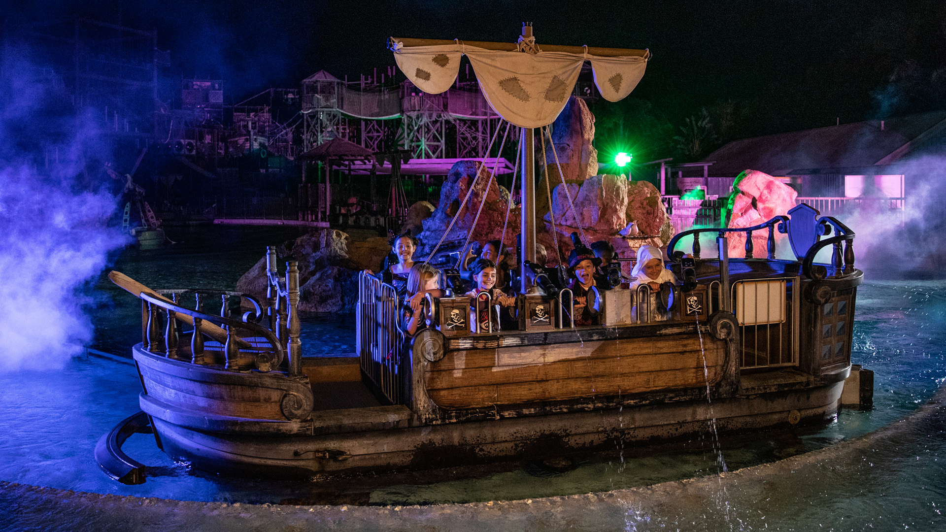 A group of people ride a pirate-themed boat through water at night, surrounded by colorful lights, mist, and rocky decorations at an amusement park or themed attraction.