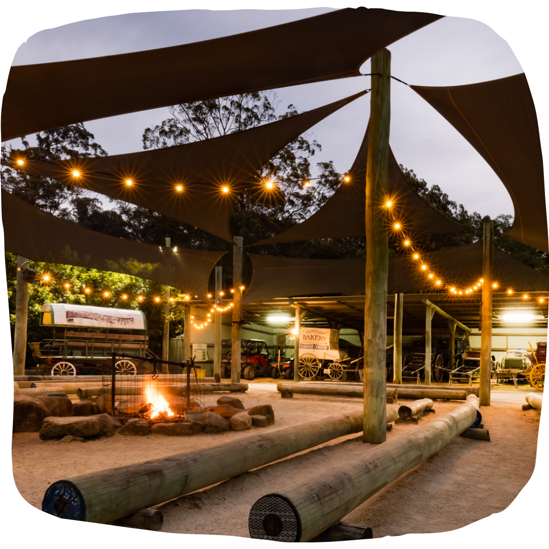 Outdoor seating area with a fire pit, string lights, wooden benches, and shade sails, surrounded by trees at dusk.