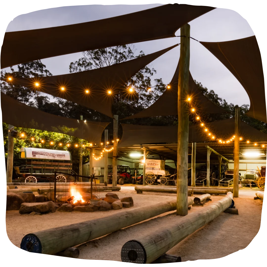 Outdoor seating area with a fire pit, string lights, wooden benches, and shade sails, surrounded by trees at dusk.