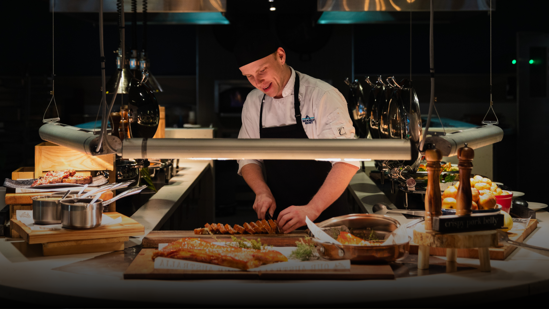 A chef in a white jacket and black apron slices food at a well-lit buffet counter, surrounded by trays of bread, dishes, and culinary utensils, with a warm, inviting ambiance.