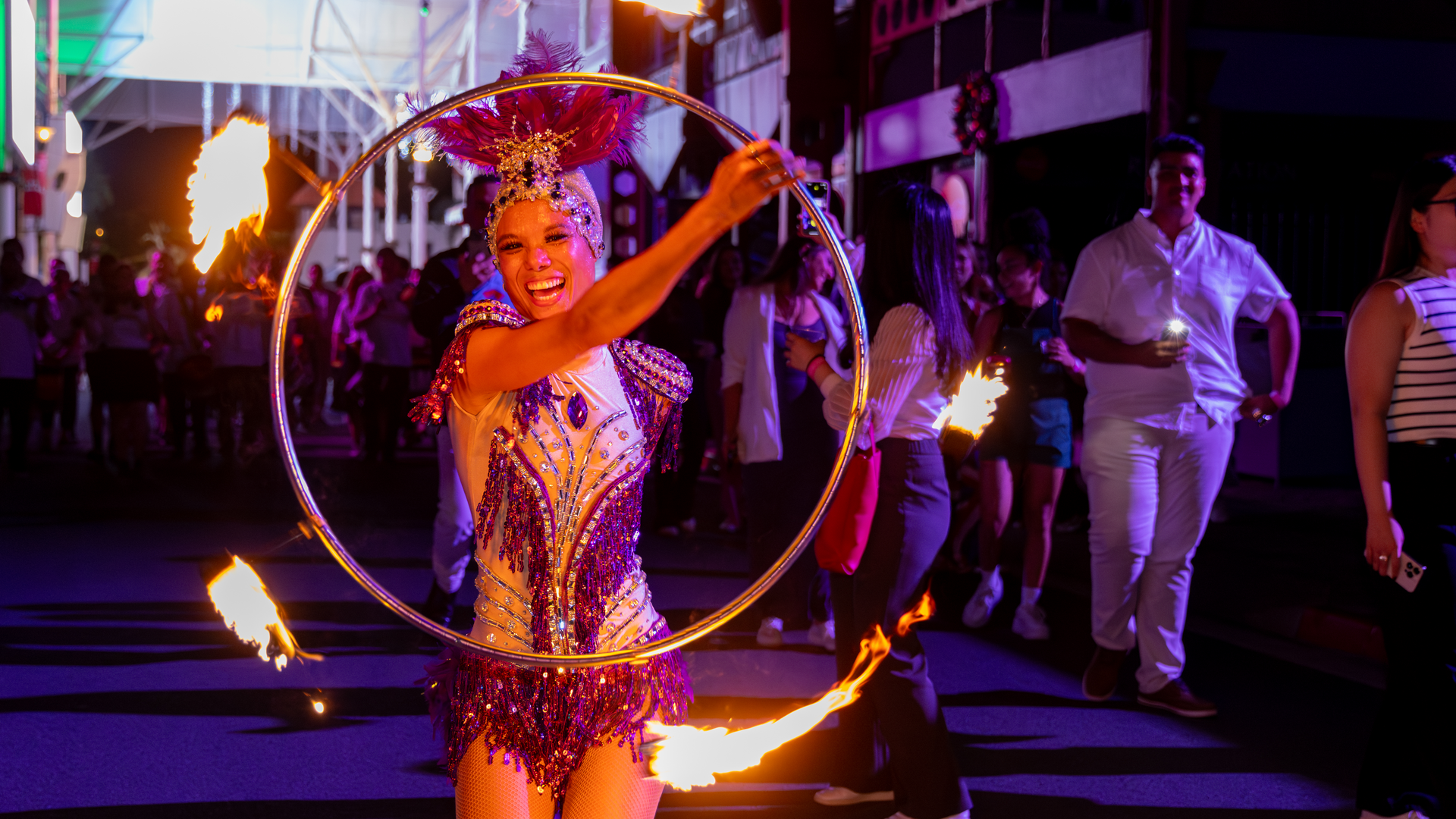 A performer in a sparkling costume and feathered headpiece hula-hoops with a ring surrounded by flames during a lively nighttime street event, with a crowd watching in the background.