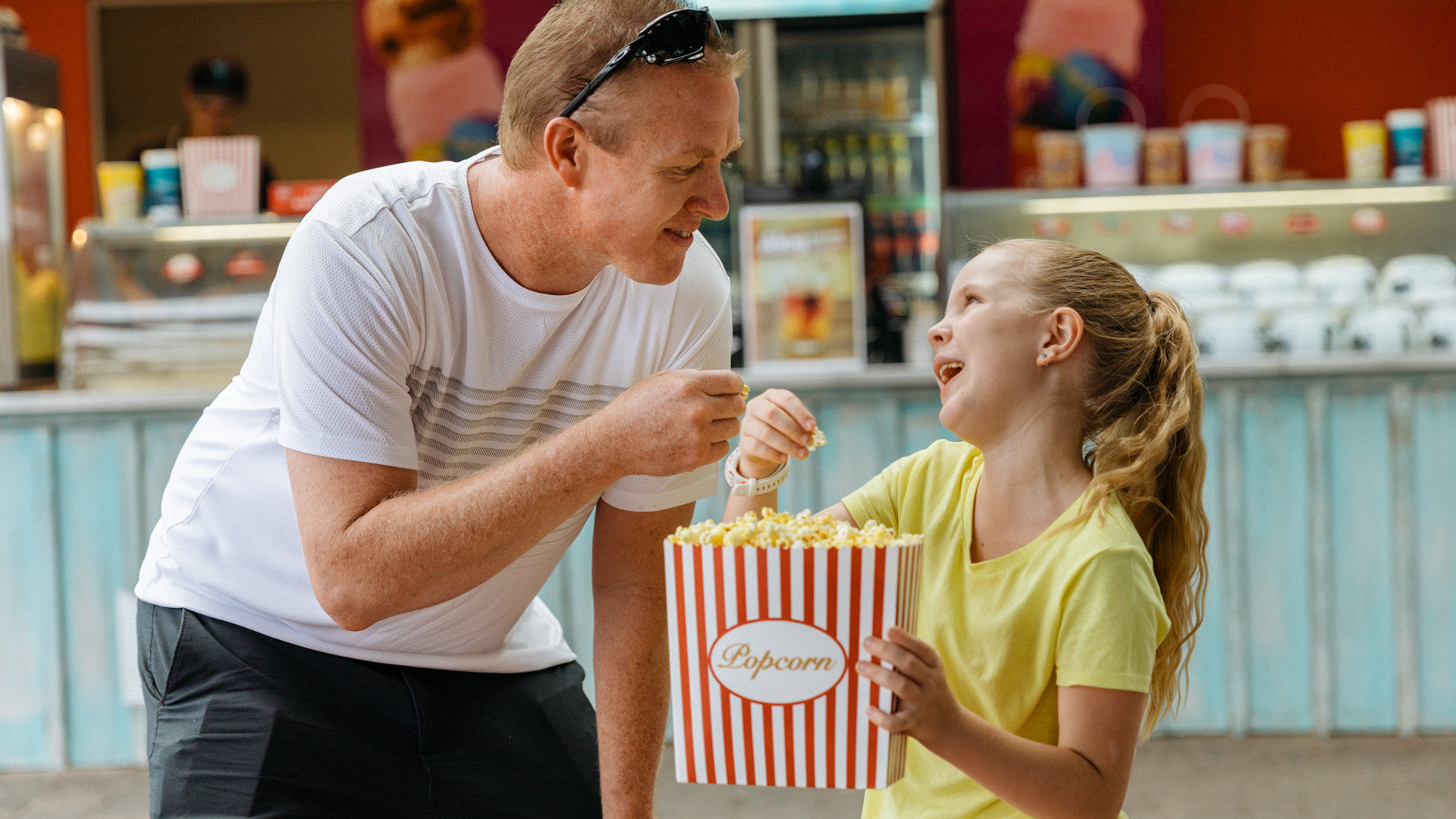 An adult and a child share a container of popcorn at an outdoor concession stand, smiling and enjoying their snack together.