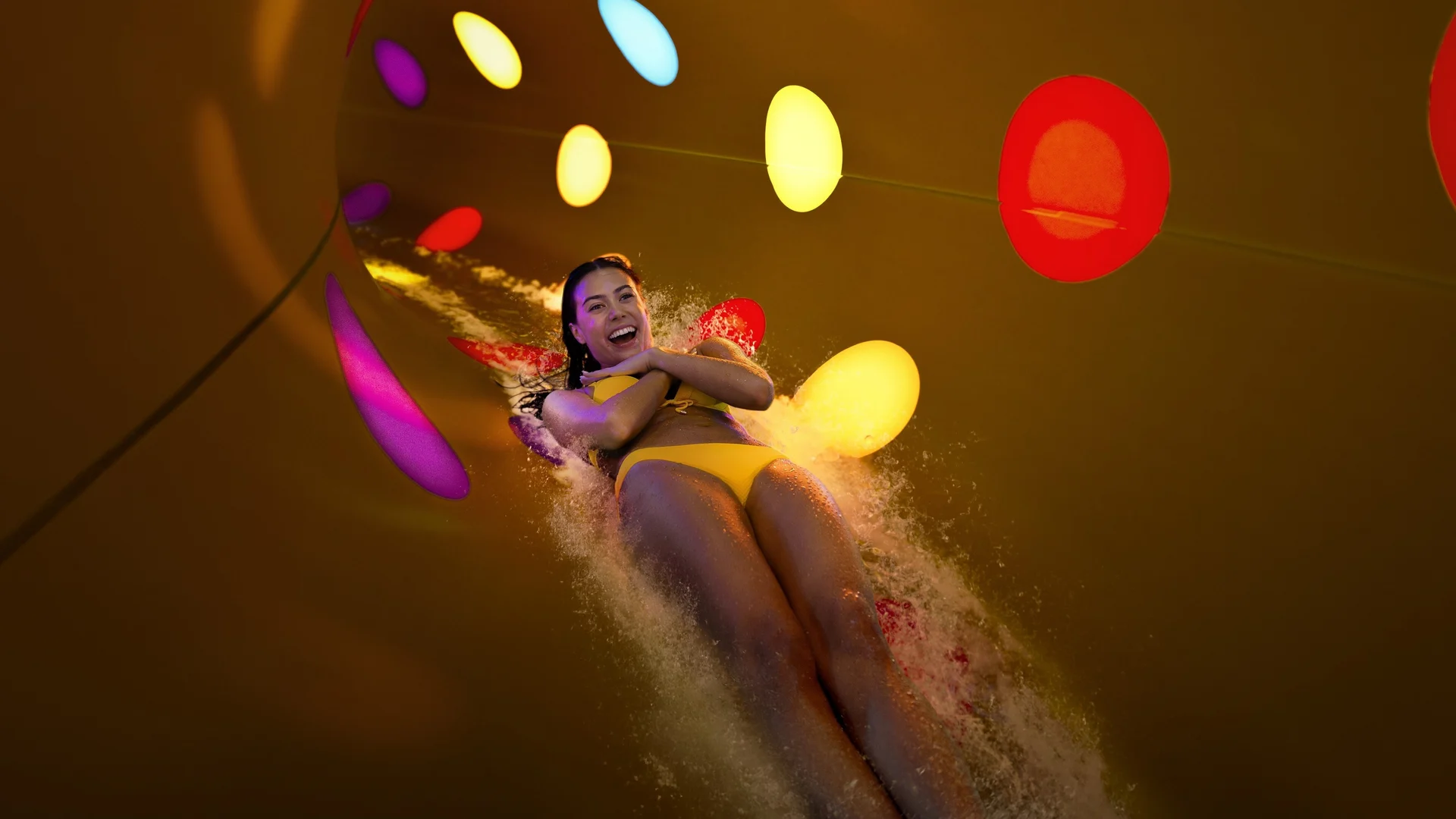 A woman in a yellow swimsuit slides down a water slide with colorful circular lights on the tunnel walls, smiling and crossing her arms over her chest as water splashes around her.