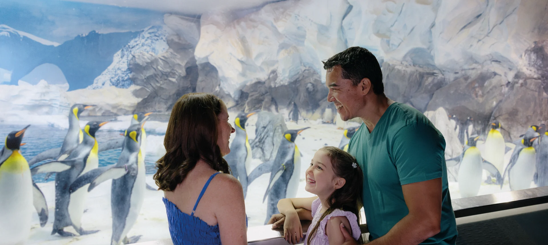 A family of three smiles and looks at each other while standing in front of a glass enclosure with many penguins and an icy, snowy backdrop at an aquarium or zoo exhibit.