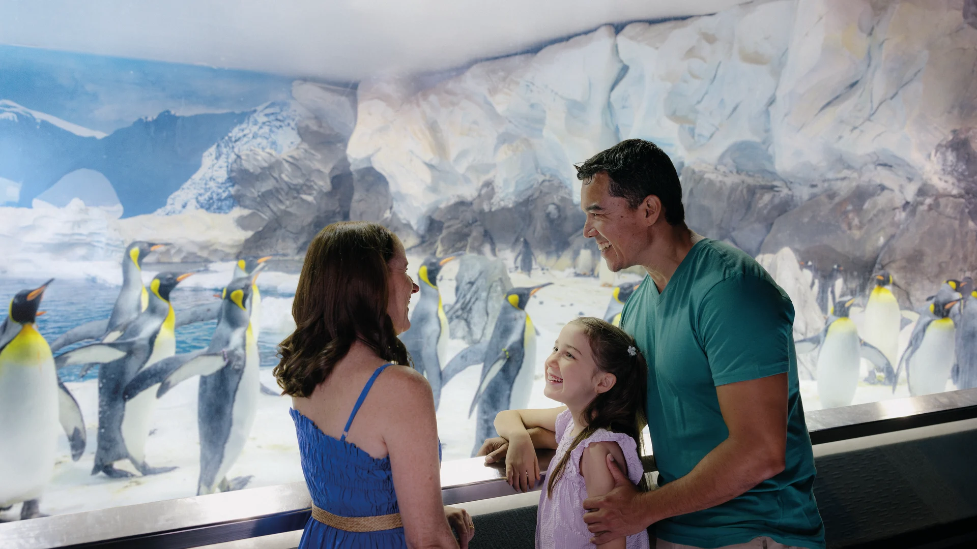 A family of three smiles and looks at each other while standing in front of a glass enclosure with many penguins and an icy, snowy backdrop at an aquarium or zoo exhibit.