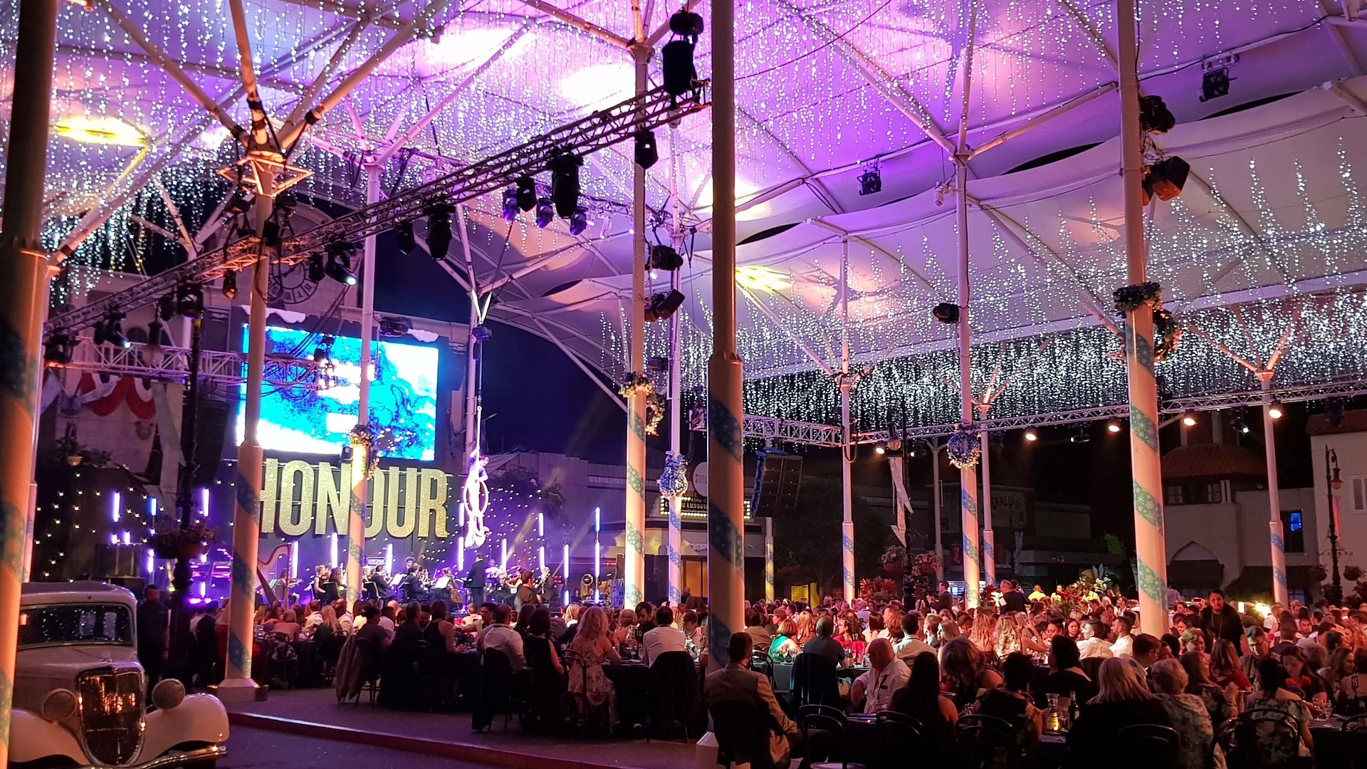 A large, festive event takes place under a canopy decorated with hanging lights. Many people are seated at tables, and a large "BONJOUR" sign and video screen are visible in the background.