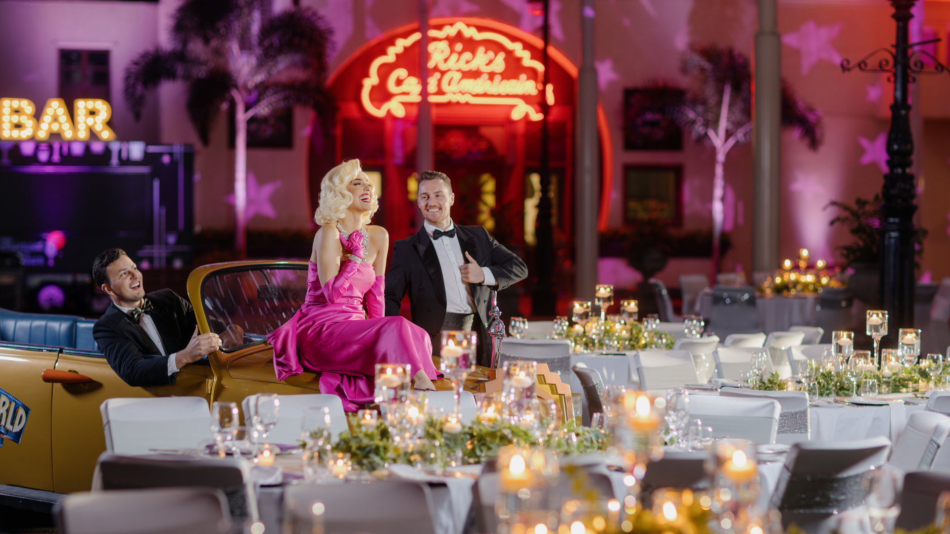 Three people dressed in formal, vintage-style clothing pose near a yellow convertible amid elegant candlelit tables. Neon signs and palm trees create a glamorous, old Hollywood atmosphere in the background.