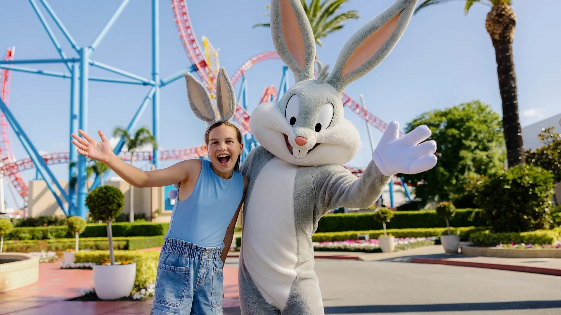 Child posing happily with a Bugs Bunny at Warner Bros. Movie World, with roller coaster tracks and palm trees in the background.