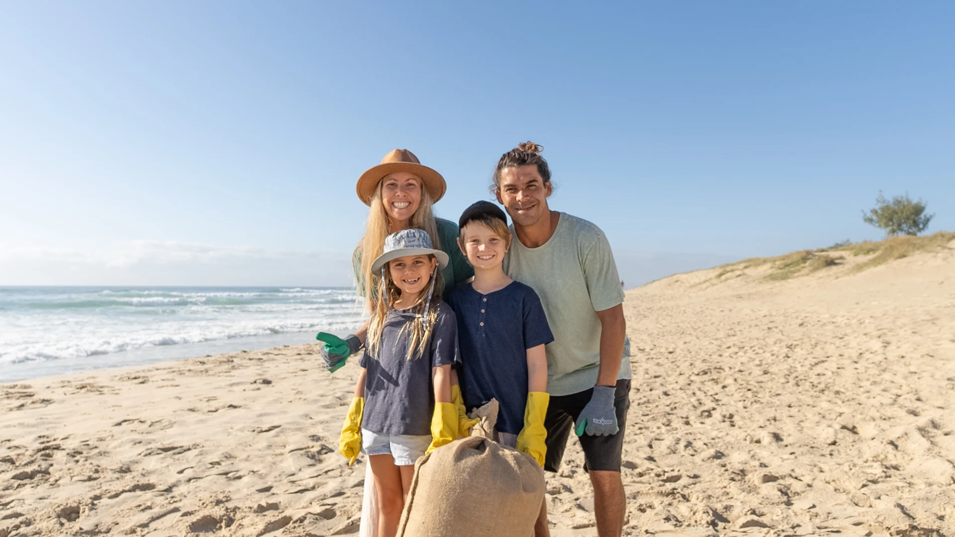 A smiling family of four stands on a sandy beach with cleaning gloves and a large burlap bag, participating in a beach cleanup on a sunny day. The ocean and dunes are visible in the background.