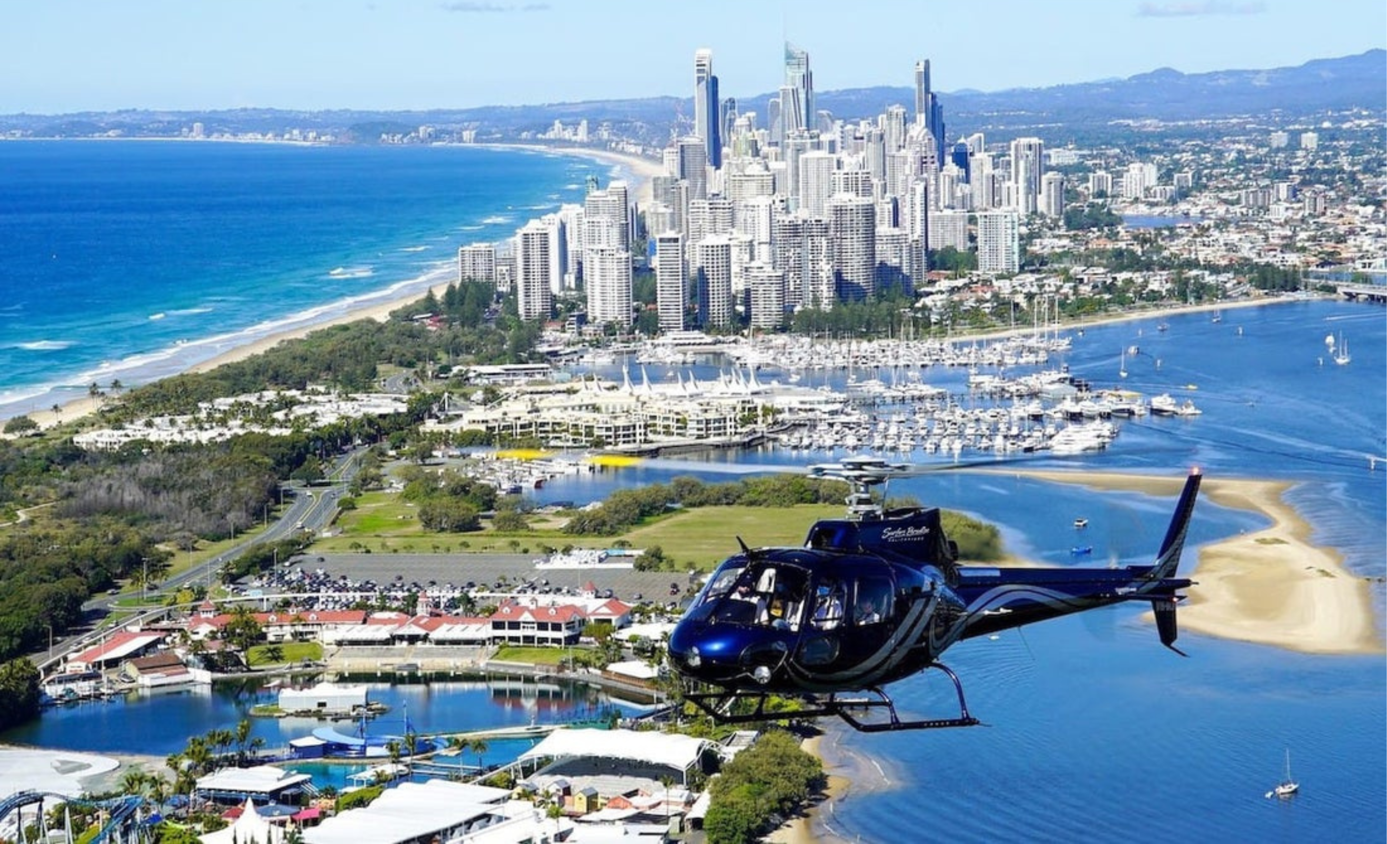 A helicopter flies over a coastal city with high-rise buildings, marinas, and a beach bordered by blue ocean and greenery under a clear sky.