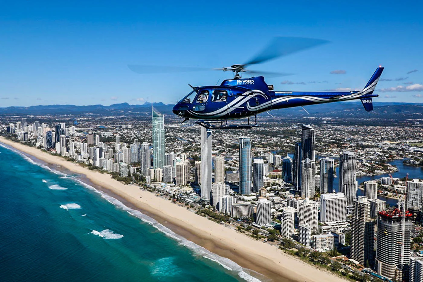 A helicopter flies over a coastal cityscape with high-rise buildings, sandy beach, and ocean waves under a clear blue sky.