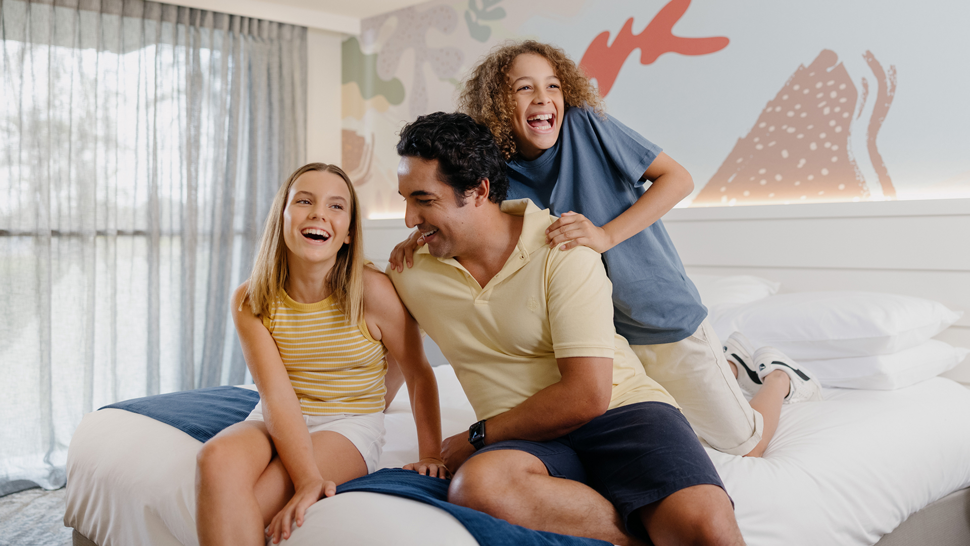 A cheerful family of three sits and laughs together on a neatly made bed in a bright, modern bedroom with large windows and colorful wall art in the background.