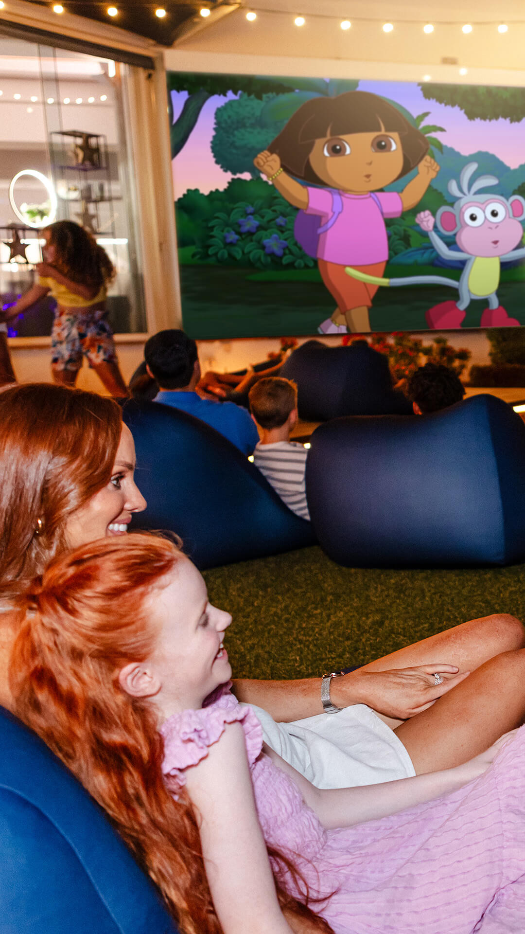 A mother and daughter, as well as families in the background, watch a Dora the Explorer movie on a big screen.