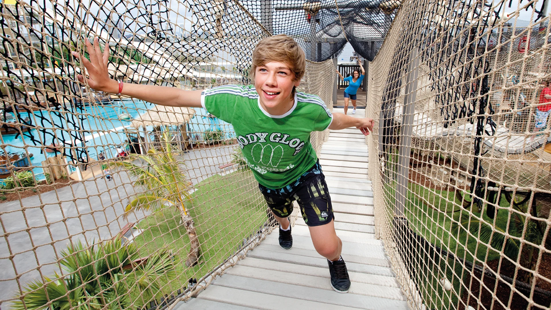 Teen boy wearing a green shirt and shorts smiles while walking on a rope bridge above an outdoor waterpark area.