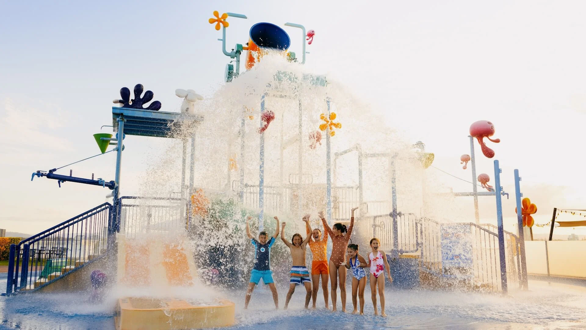 Children play under a large splash of water at a colorful outdoor water park structure, with slides and decorations on a sunny day.