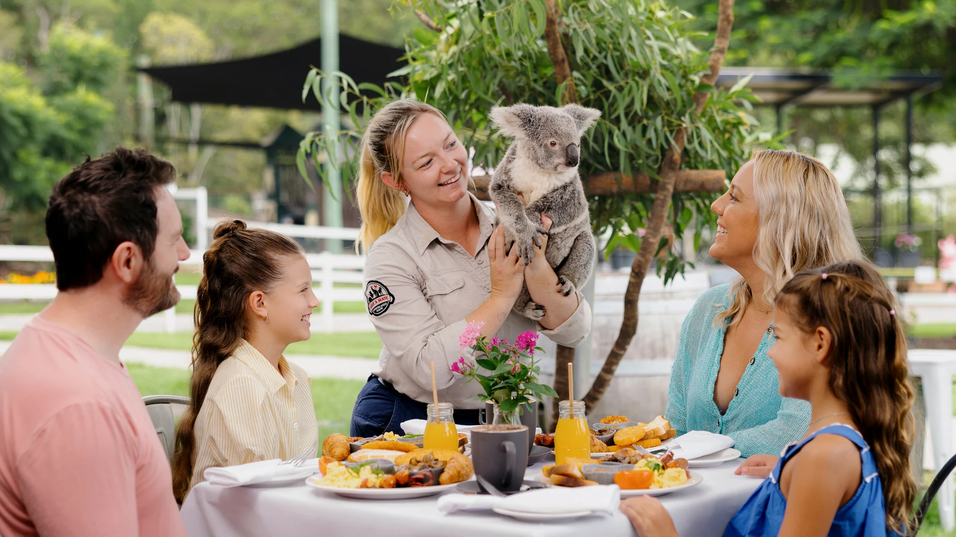 A woman holding a koala stands by a table outdoors, smiling at a family of four enjoying a meal together. The setting is lush and green, with trees and plants in the background.